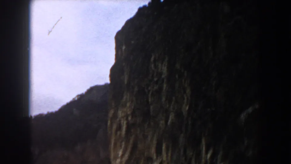 1960 Italy 8mm frame with a shadowed rock face rising in front of pale sky and a lower coastal hillside.
