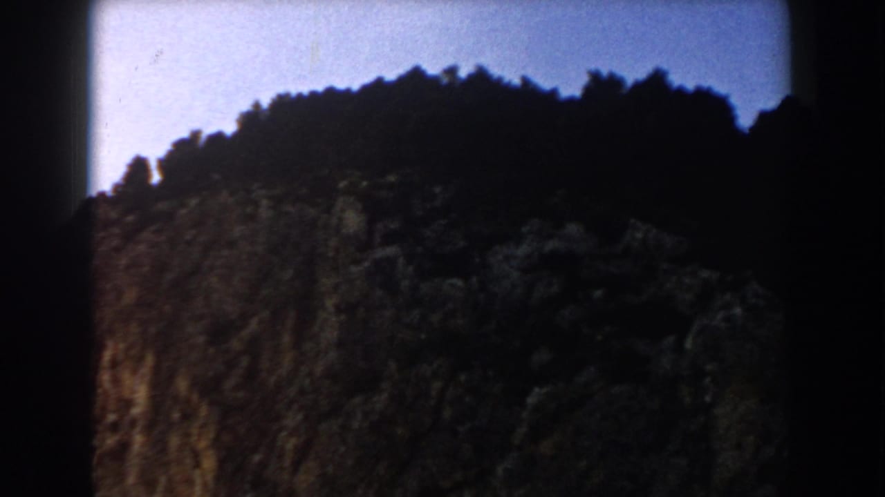 1960 8mm Italy landscape frame with a forested cliff top, exposed rock, and blue sky.