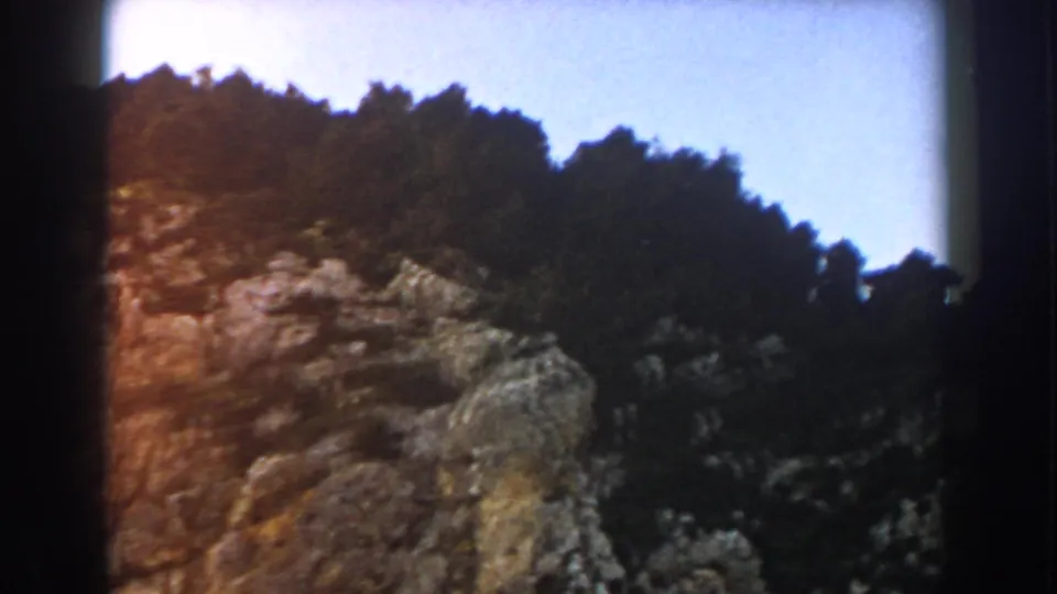1960 8mm Italy travel frame showing jagged rock, dense trees, and warm film flare near the edge.