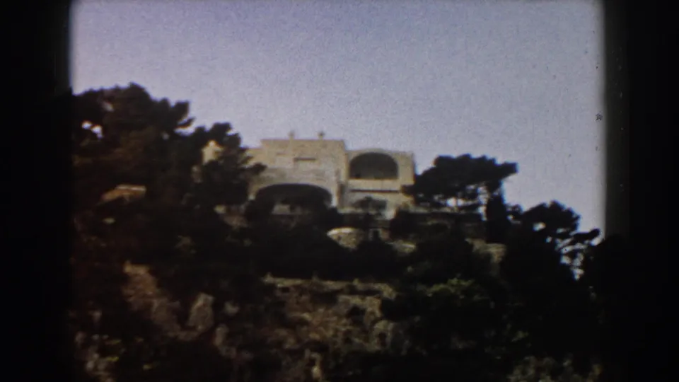 1960 Italian coastal view of a white building set above trees on a rocky slope, with blue sea visible behind it.