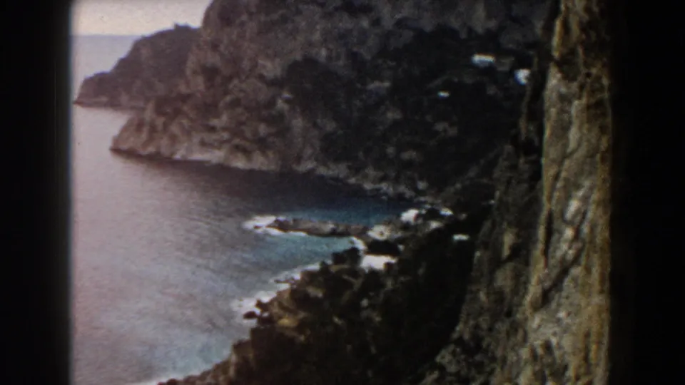 1960 8mm Italy footage looking down along a steep rocky coast toward blue water and a small pale shoreline.