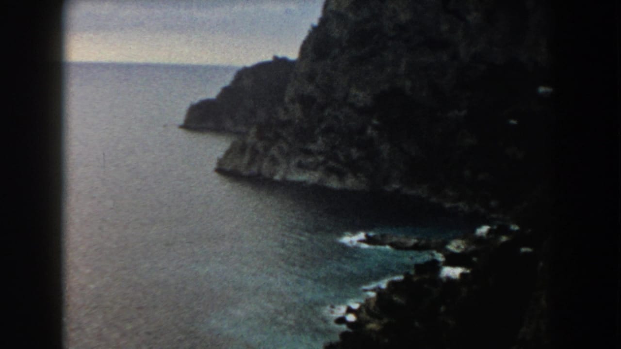 Dark rocky cliff above calm sea with shoreline surf below.