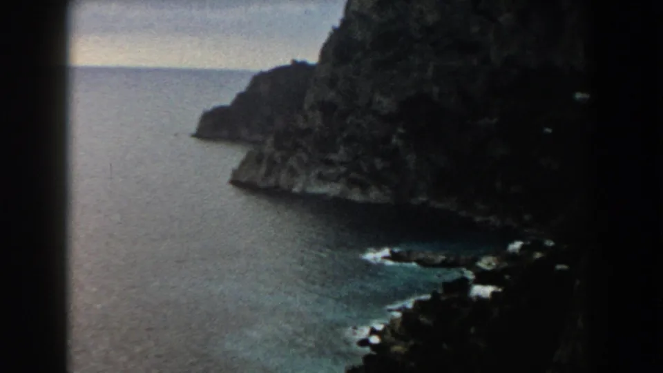 1960 Italy 8mm view of a steep rocky coastline meeting the sea, with dark cliff faces and small waves below.