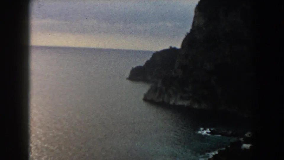 1960 Italy 8mm frame of a rocky headland beside open water, partially framed by dark film gate edges.