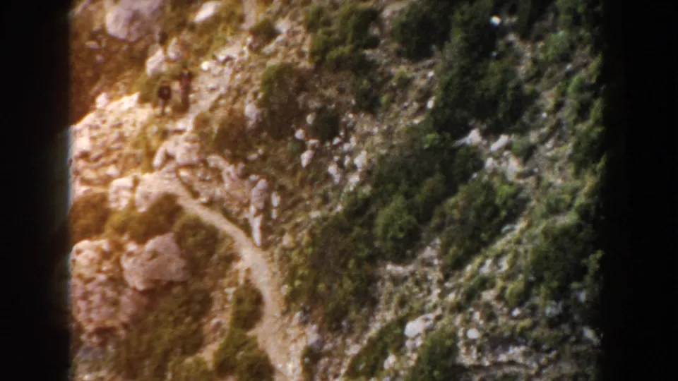 1960 Italy 8mm view of rough coastal terrain with a winding path, scrub, boulders, and small distant visitors.