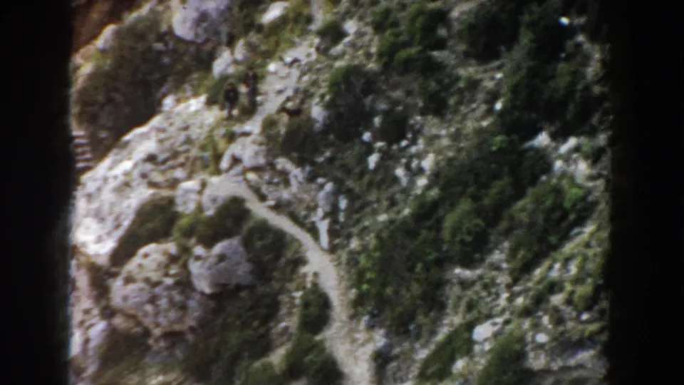 1960 Italy 8mm frame showing a scrub-covered rocky slope, footpath, boulders, and distant people on the terrain.
