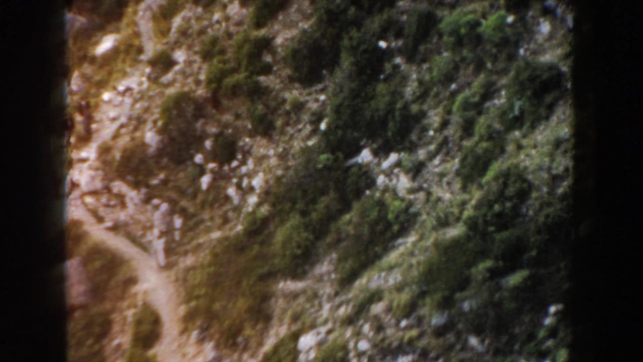 A narrow mountain trail appears among rocks, scrub, and small trees in 1960 Italy home-movie footage.