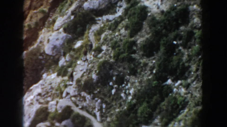 Distant hikers continue upward on a rugged Italian mountain path, seen in grainy 1960 8mm footage.