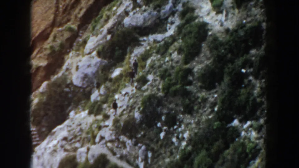1960 home-movie frame from Italy with small male hikers moving through a rocky, shrub-covered mountain slope.