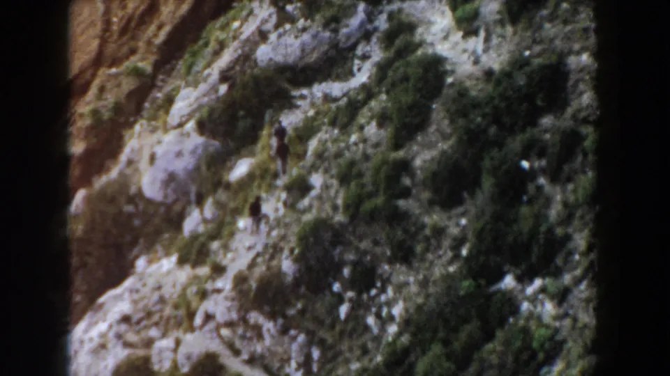 Final 1960 Italy 8mm frame showing distant men ascending a narrow path through rugged rocks and mountain vegetation.