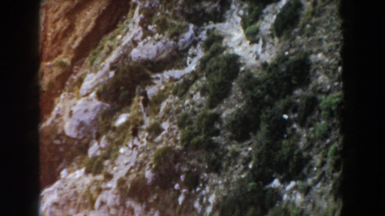 Rocky slope with shrubs and a small distant hiker near a narrow path.