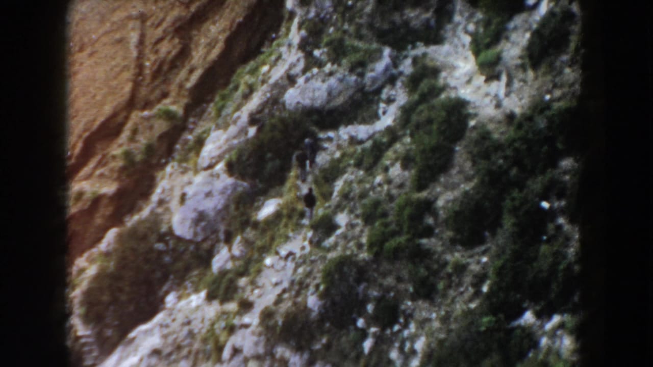 1960 Italy 8mm frame from above, with a small hiker on a stony hillside among shrubs and exposed rock.
