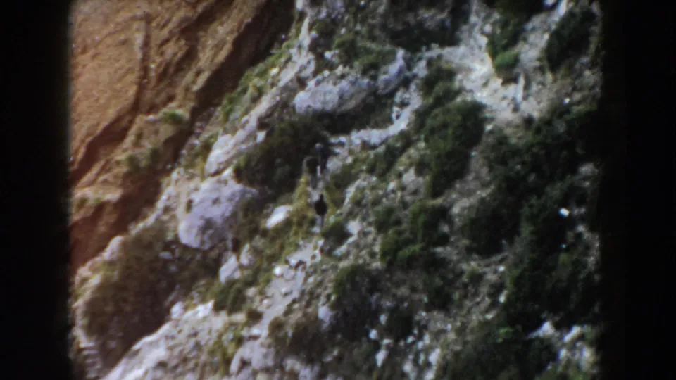1960 Italy 8mm frame from above, with a small hiker on a stony hillside among shrubs and exposed rock.