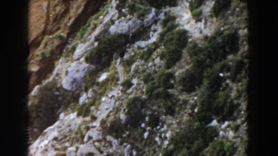 1960 Italy 8mm frame showing a distant hiker crossing a rugged hillside with shrubs, stones, and dark gate borders.