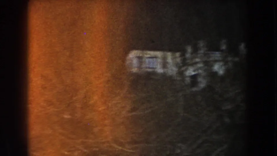 Soft 8mm view in Italy of a pale camper-like structure partly obscured by trees and heavy warm flare.