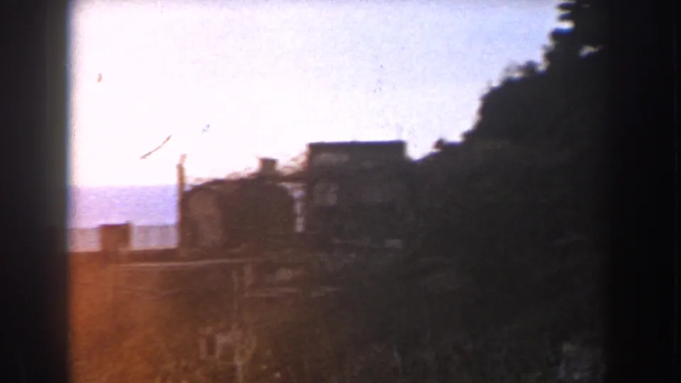 1960 Italy home-movie frame with rooftops, a dark tree-covered edge, pale sky, and distant horizon.