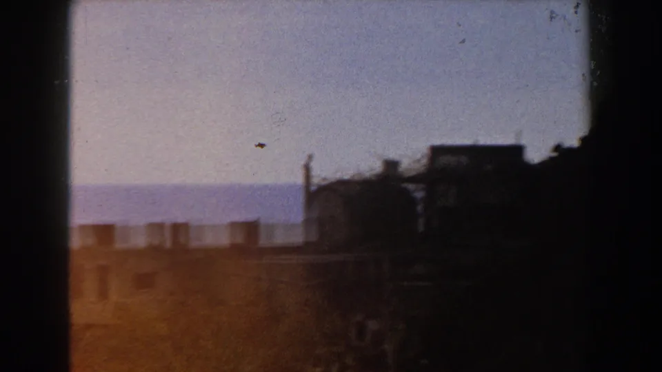 Soft 8mm Italy frame showing rooftop silhouettes, a pale sky, distant blue horizon, and dark tree mass.