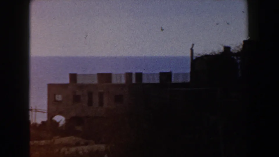 1960 Italy 8mm travel frame showing a shadowed crenellated castle wall with arched openings near a hazy sea horizon.