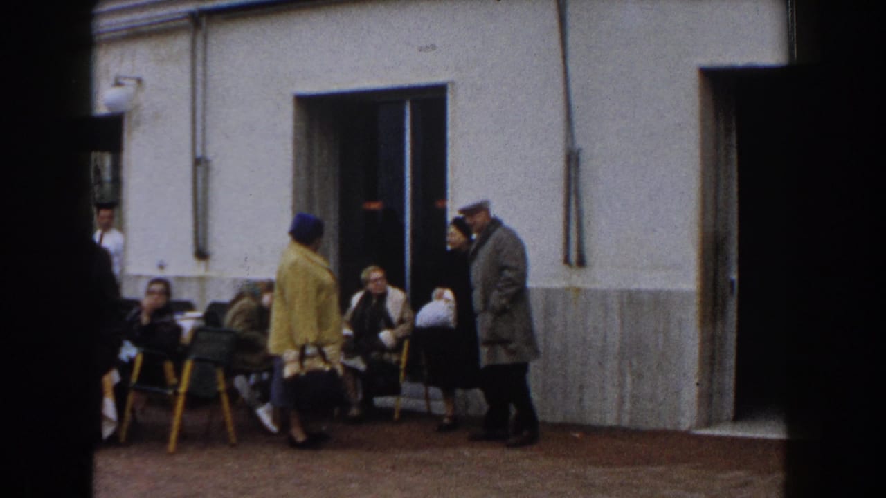 Older adults gather near a white stucco doorway with yellow chairs and outdoor tables.