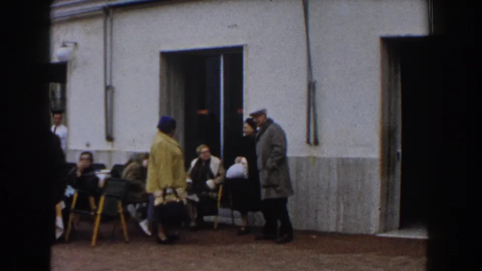 In 1960 Italy home-movie footage, older adults stand and sit outside a stucco cafe-like entrance with yellow chairs nearby.