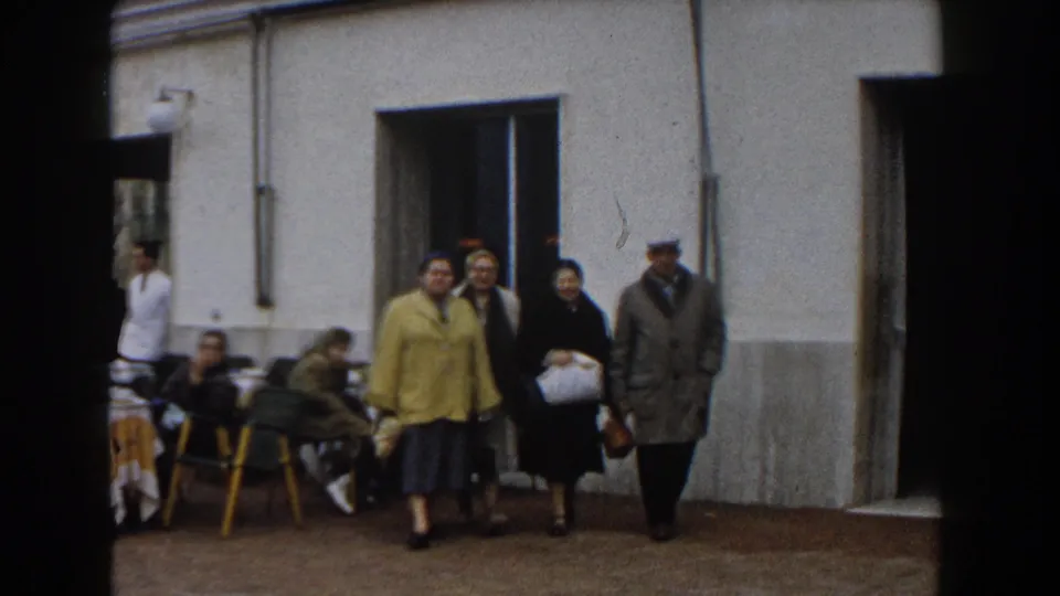 Several older adults, including a woman in a yellow coat, walk toward the camera in 1960 Italy home-movie footage.