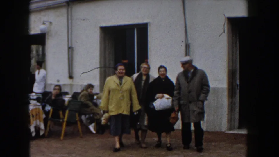 Four older adults walk together outside a cafe-like entrance, with seated people and yellow chairs behind them in Italy.