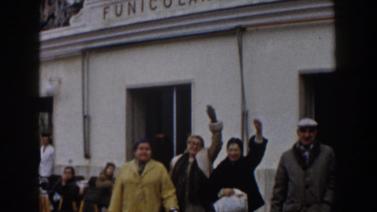 1960 Italy frame with several adults waving toward the camera in front of a building marked Funicolare, with seated people in the background.