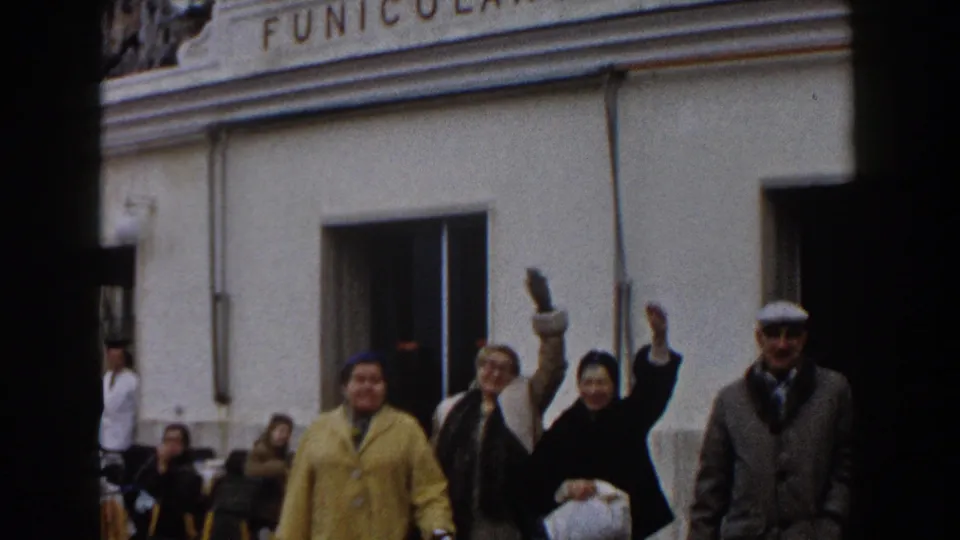 1960 Italy frame with several adults waving toward the camera in front of a building marked Funicolare, with seated people in the background.