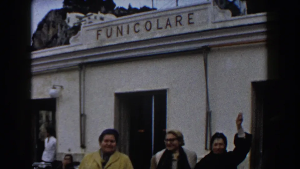 Closer 1960 Italy home-movie view of adults near the funicolare facade, including a yellow coat and a raised hand greeting the camera.