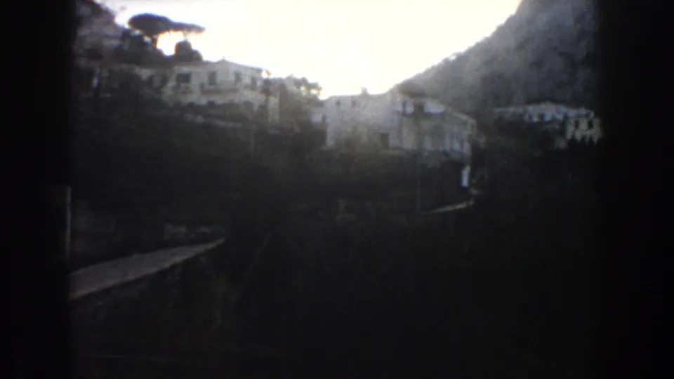 Underexposed 1960 Italy home-movie frame showing hillside buildings, a road or track, and strong dark film edges after the station view.
