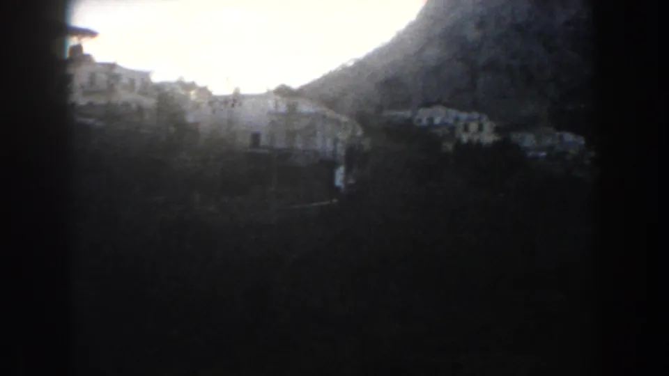 1960 8mm home-movie view in Italy showing pale hillside houses beneath a dark mountain, partly obscured by heavy foreground shadow.