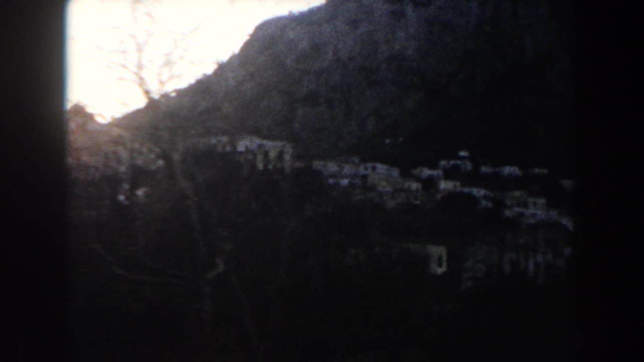 Shadowed 1960 8mm view across an Italian mountain village, with small light-colored buildings scattered along the slope.