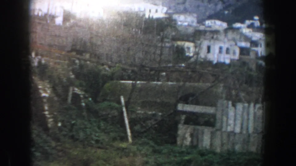 Closer 1960 8mm view in Italy showing hillside houses, terraced ground, overgrown vegetation, and rough wooden fencing.