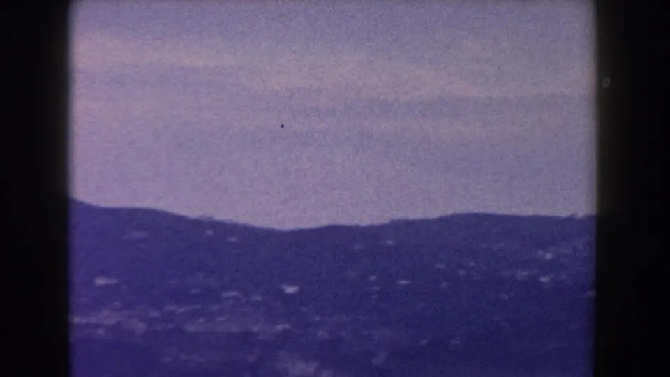 The pan opens to a distant hillside in Italy, with small light buildings scattered across the slope beneath a hazy sky.