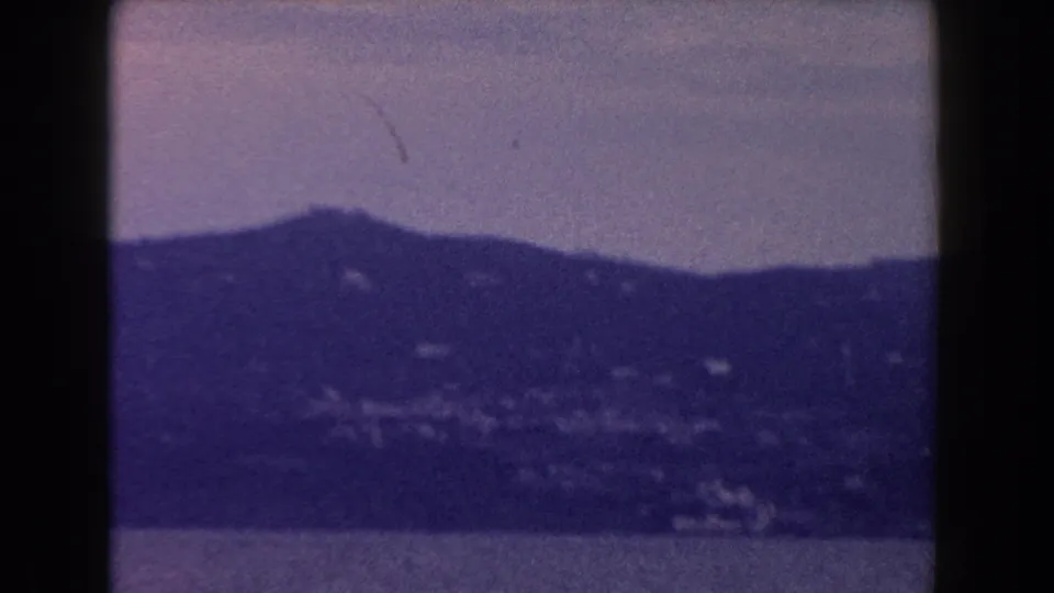 A band of sea sits below distant Italian hills dotted with small buildings, rendered in soft grainy 8mm color.