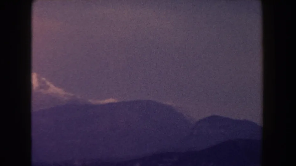 1960 Italy 8mm frame showing a snow-covered mountain peak beyond dark ridges in hazy afternoon-like light.