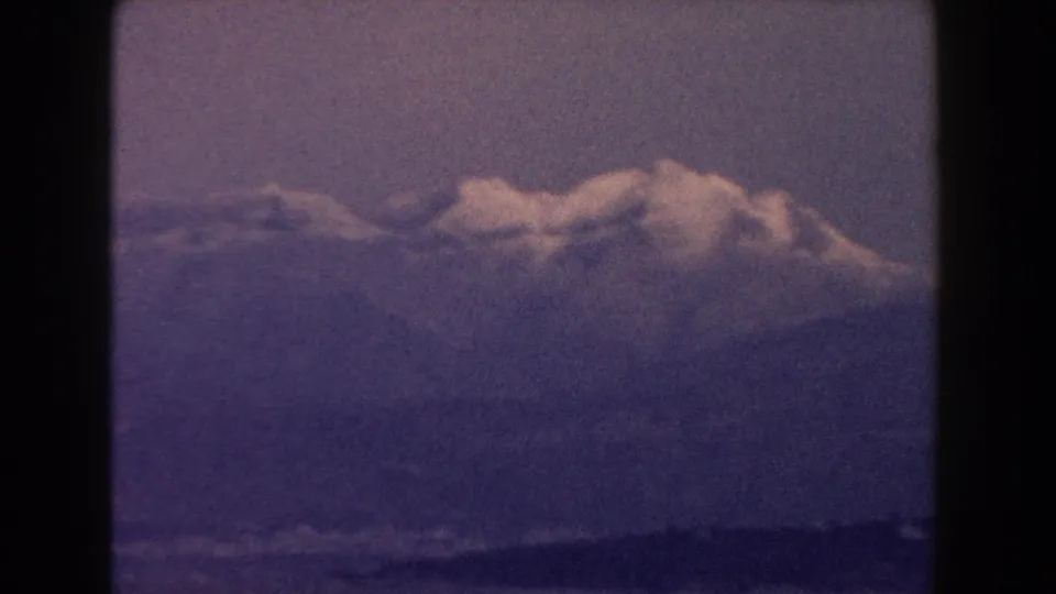 1960 Italy 8mm frame showing a distant snow-capped mountain ridge over dark water under a hazy purple sky.
