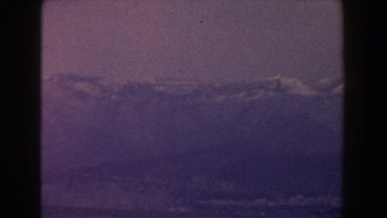 1960 Italy 8mm frame with a clouded mountain range and dark water below, tinted by aged magenta-blue film color.