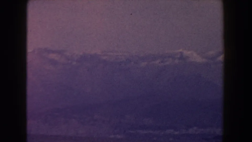 1960 Italy 8mm frame with a clouded mountain range and dark water below, tinted by aged magenta-blue film color.