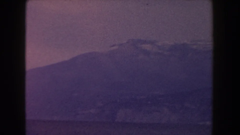 1960 Italy 8mm frame showing a dark mountain slope, summit cloud, and foreground water through faded purple film stock.