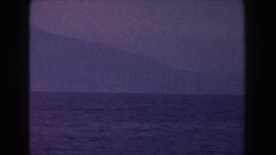 1960 Italy 8mm frame with dark open water and a faint mountain form under a hazy, purple-blue sky.