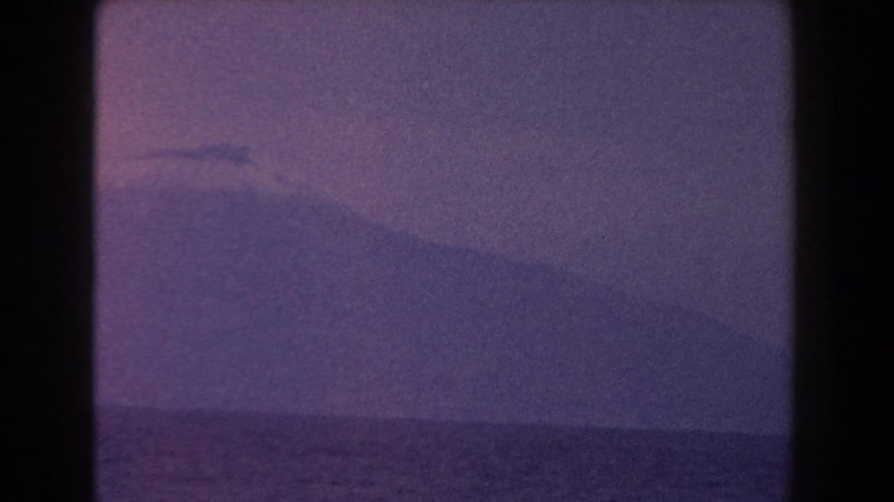 1960 Italy 8mm frame with a hazy mountain rising beyond the sea and a faint mist or cloud near its summit.