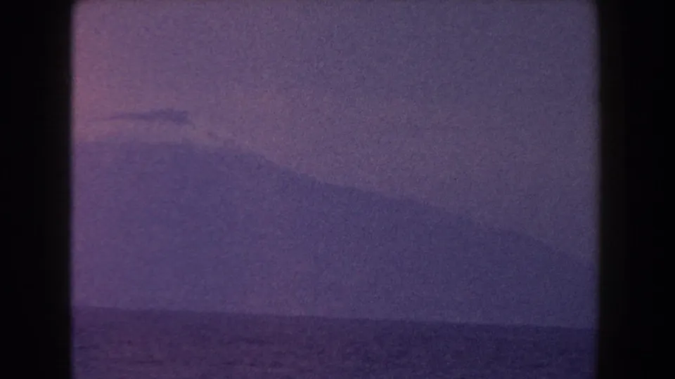 1960 Italy 8mm frame with a hazy mountain rising beyond the sea and a faint mist or cloud near its summit.