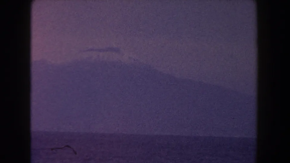 1960 Italy 8mm frame showing the sea and a distant mountain softened by haze, grain, and low-contrast exposure.