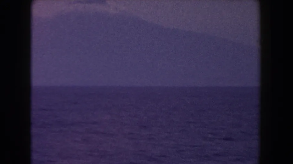 1960 Italy 8mm frame holding on open water and a distant mountain form under muted blue-purple light.