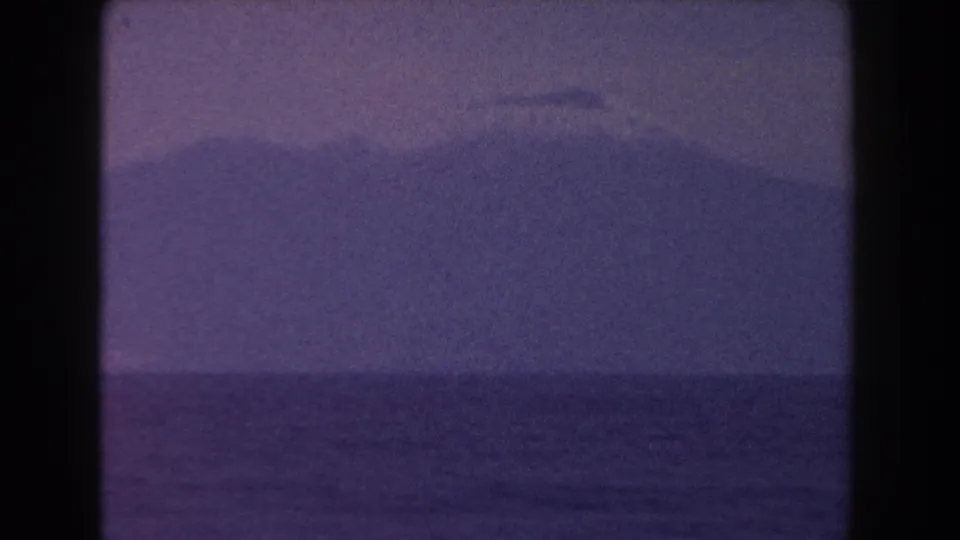 1960 Italian 8mm frame showing a distant mountain silhouette beyond calm water under a faded purple-blue sky.