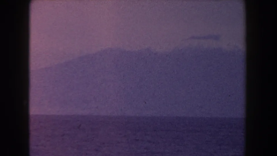 1960 Italian 8mm frame showing a soft mountain silhouette past open water, with muted magenta and blue film tones.