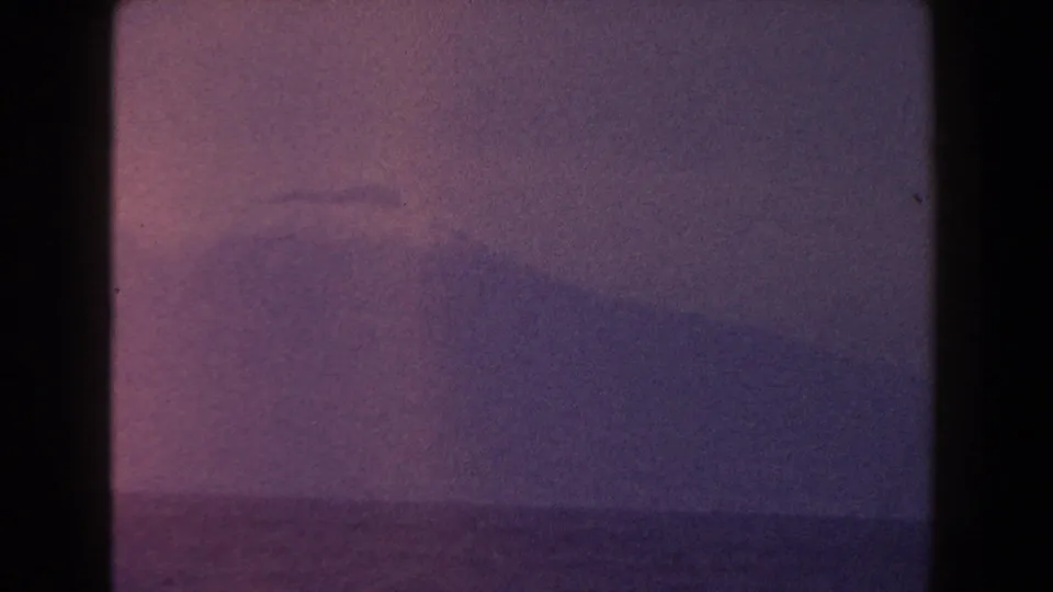1960 Italian 8mm frame with a distant coastal mountain, low ocean horizon, and faded purple-pink twilight tones.