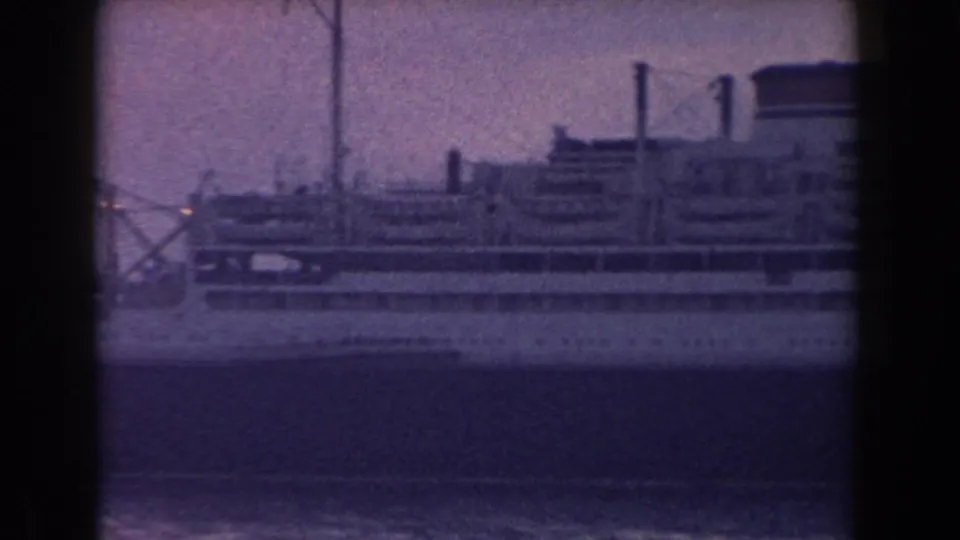1960 Italian 8mm frame showing the side of a large docked ship with deck levels, funnels, and soft coastal haze.