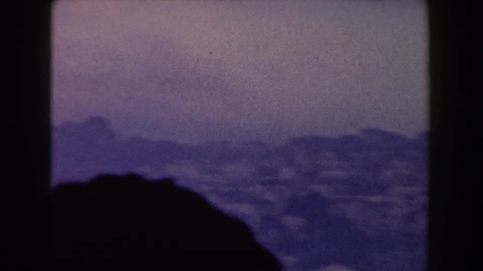 1960 Italy home-movie frame showing a hazy waterfront city or harbor area below hills, partly blocked by a dark foreground silhouette.
